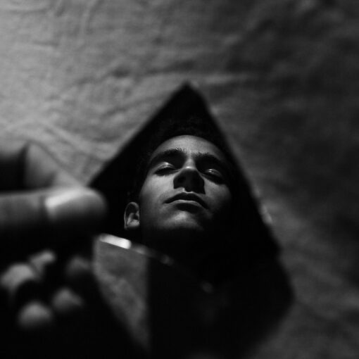 Das Gesicht einer Person wird in einem kleinen, unregelmäßig geformten Spiegel reflektiert, der von einer Hand gegen eine strukturierte Oberfläche gehalten wird, in Schwarz und Weiß. (KI generierte Beschreibung)