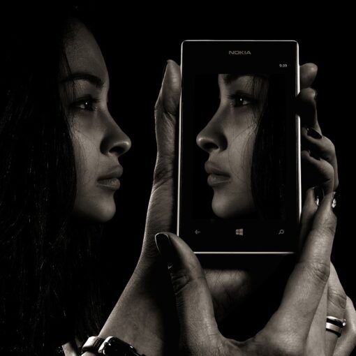 Eine Person hält ein Smartphone in der Hand, das ihr Profil zeigt und ihr Gesicht spiegelt, wobei sowohl das Telefon als auch das Gesicht in Graustufen vor einem schwarzen Hintergrund dargestellt sind. (KI generierte Beschreibung)