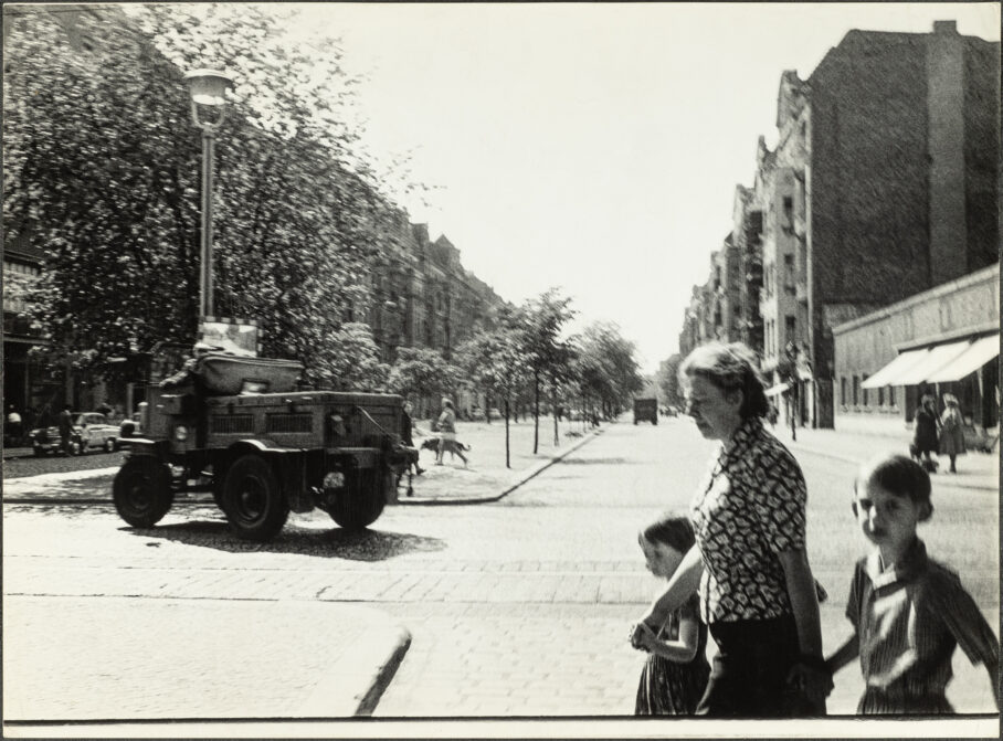 eine Frau hat zwei Kinder an der Hand, die Straßenflucht ist sonnenbeschienen, die Bäume jung, ein Militärfahrzeug biegt ab