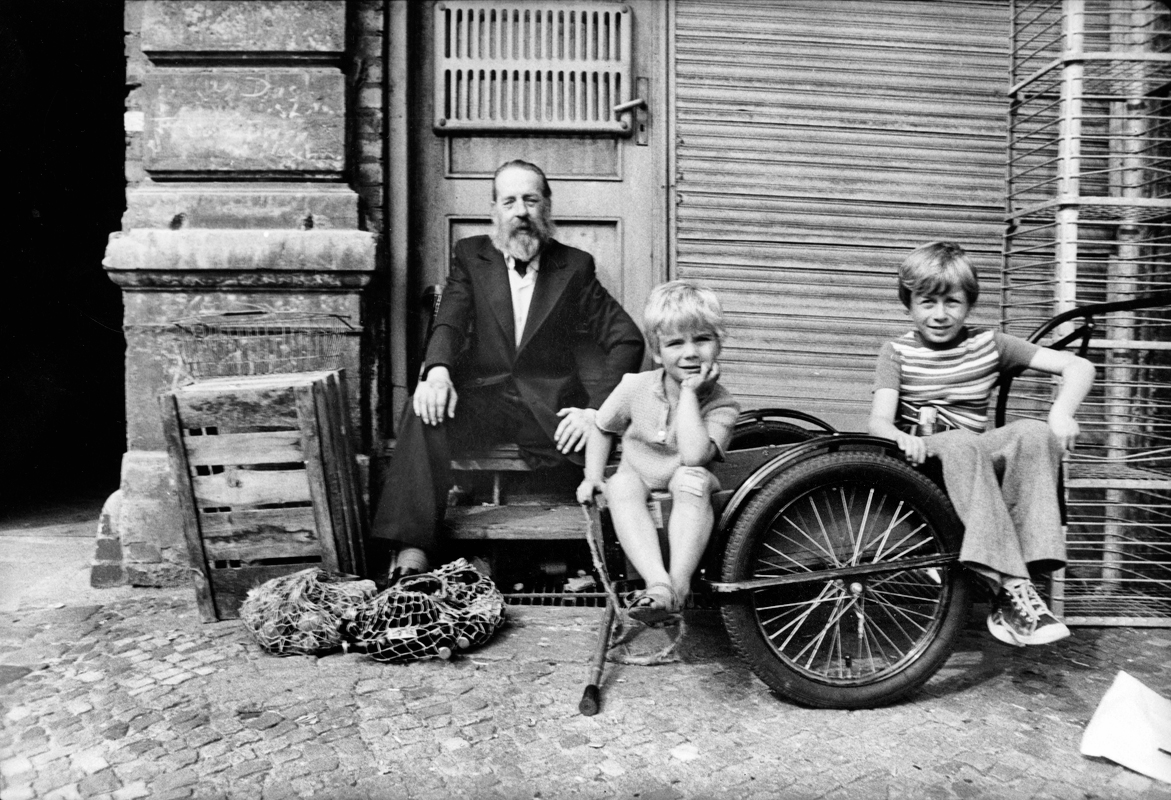 ein Mann im Anzug sitzt neben einem geschlossenen Geschäft, vor sich ein Beutel mit Bierflaschen und zwei kleine Jungs in einem Fahrradwagen