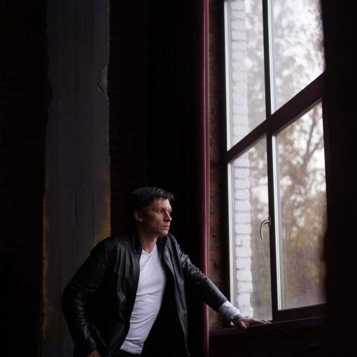 Ein Mann in schwarzer Jacke und weißem Hemd steht an einem großen Fenster und schaut mit einer Hand auf dem Fenstersims nach draußen. (KI generierte Beschreibung)