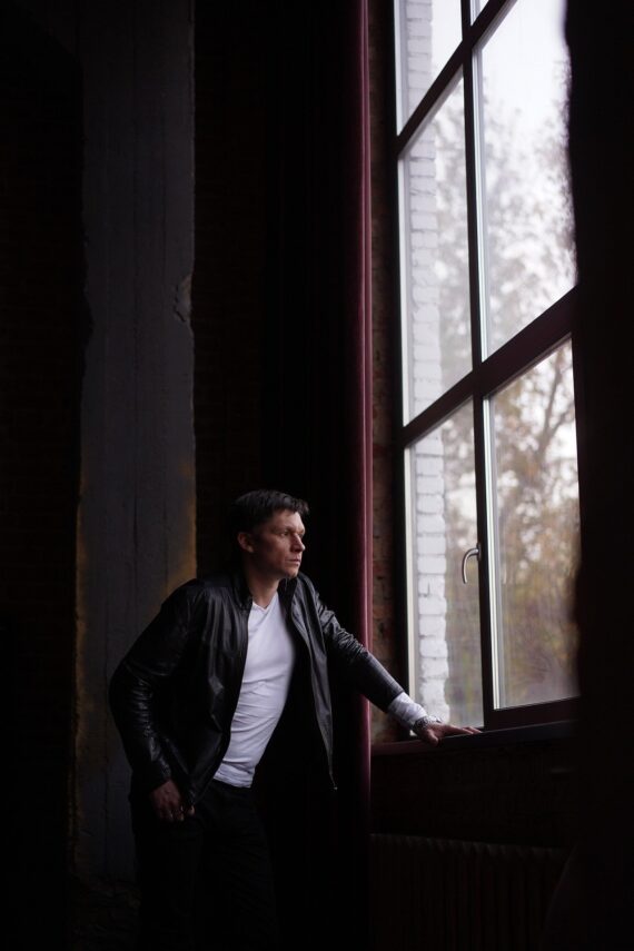 Ein Mann in schwarzer Jacke und weißem Hemd steht an einem großen Fenster und schaut mit einer Hand auf dem Fenstersims nach draußen. (KI generierte Beschreibung)