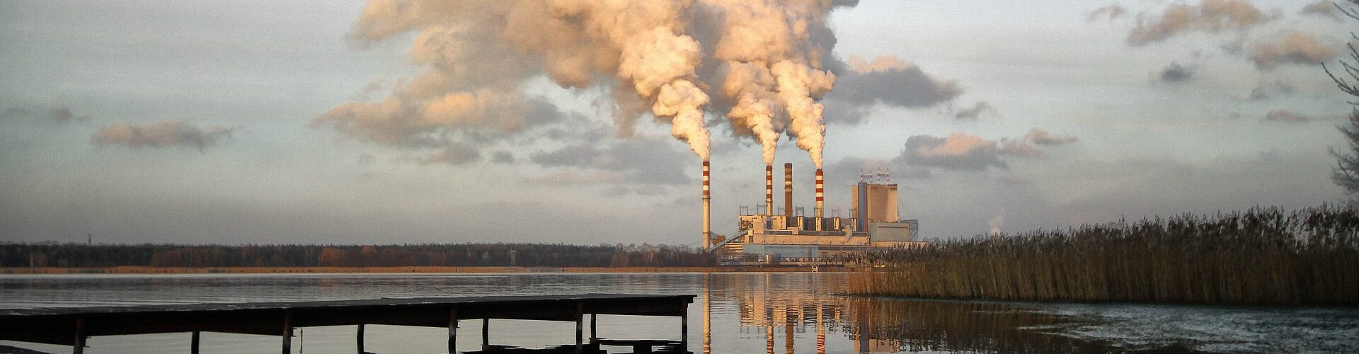 Ein Kraftwerk mit vier großen Schornsteinen stößt dicke Rauchschwaden aus, die sich in einem ruhigen Gewässer mit einem Holzsteg im Vordergrund spiegeln. (KI generierte Beschreibung)