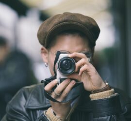 Person mit brauner Mütze und schwarzer Lederjacke, die eine Kamera an ihr Gesicht hält und ein Foto macht. Der Hintergrund ist unscharf. (KI generierte Beschreibung)