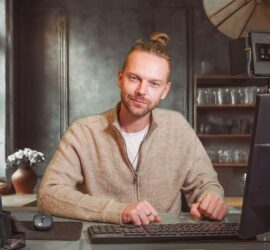 Ein Mann mit Bart und Haarknoten sitzt an einem Schreibtisch mit Tastatur und Maus und blickt in einem warm beleuchteten Raum mit Regalen und Blumen im Hintergrund in die Kamera. (KI generierte Beschreibung)